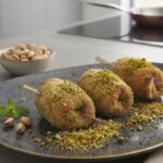 Un piatto moderno e curato presenta tre involtini di lonza impanati al pistacchio su un piano da cucina contemporaneo. Gli involtini sono dorati, croccanti e completamente chiusi e sigillati, trattenendo il ripieno all'interno. Sono guarniti con granella e farina di pistacchio, oltre a pistacchi interi e una foglia di menta. Lo sfondo è una cucina minimalista grigia con una pentola su un piano cottura a induzione e ciotole di pistacchi.