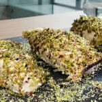 Tre involtini di pesce dalla spessa crosta croccante di granella di pistacchio verde brillante, disposti su un piatto di ardesia scuro, accompagnati da timo e limone arrostito su un tavolo di legno.